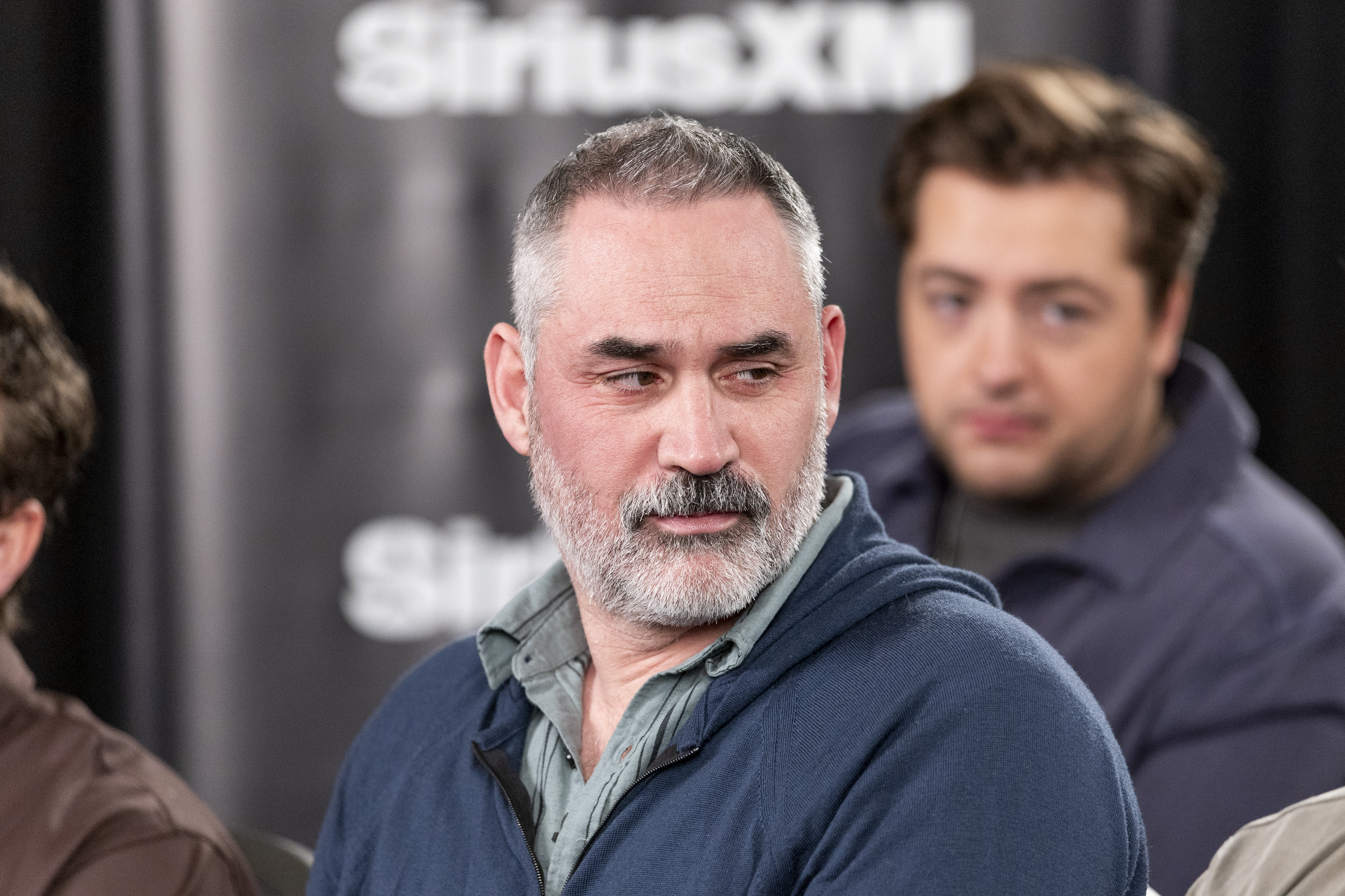 Man with short gray hair and a gray beard wearing a navy zip-up hoodie seated at a SiriusXM event, with another man blurred in the background.